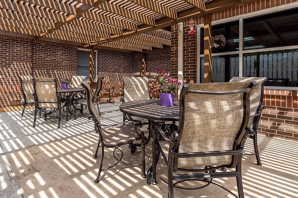 Shaded outdoor patio with metal tables and wicker-backed chairs under a wooden pergola casting striped shadows.