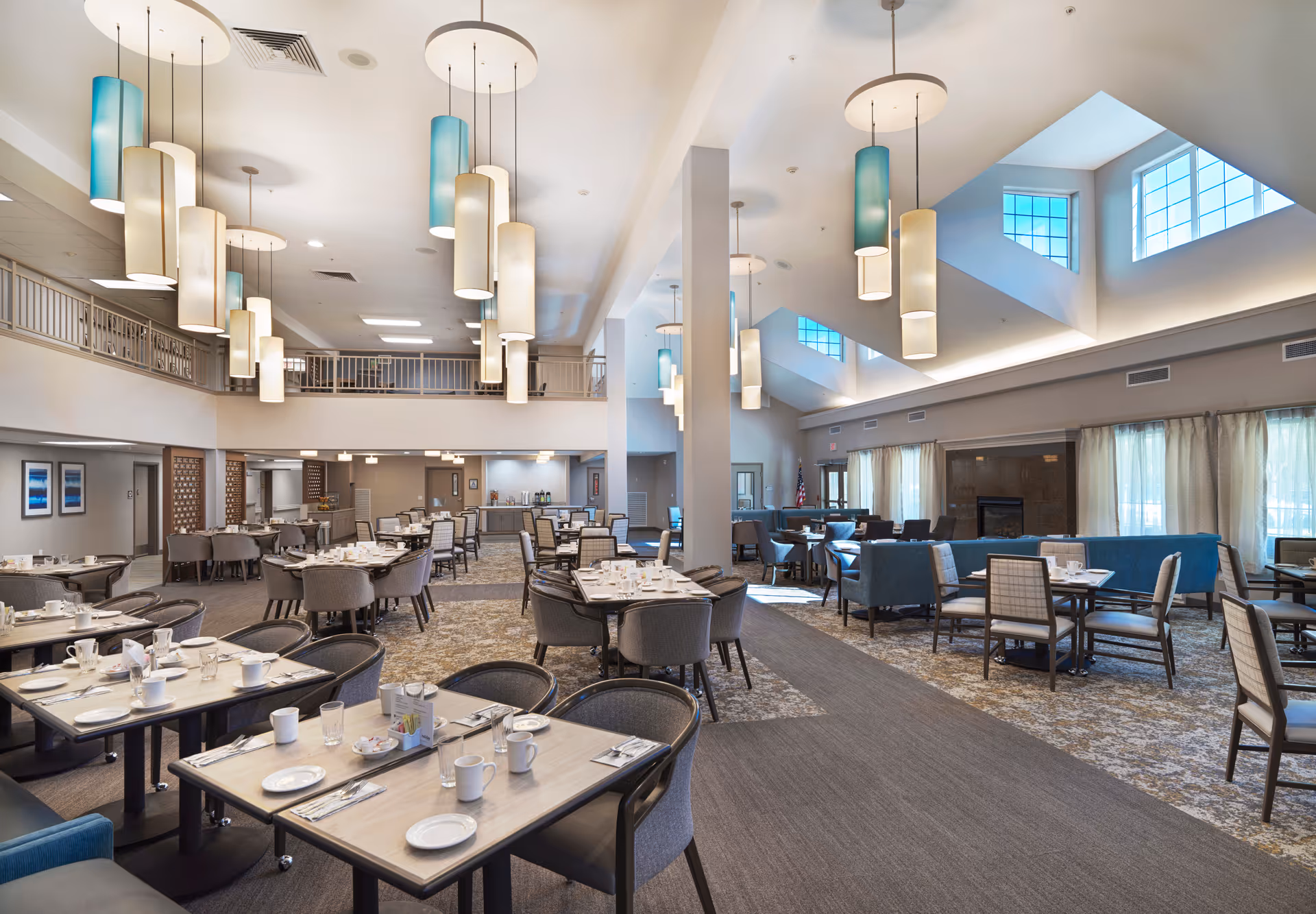 Spacious dining room in a senior living facility with multiple tables set with cups, plates, and utensils. The room features modern hanging light fixtures, large windows allowing natural light, and a mix of chairs and booth seating. The decor includes neutral tones with blue accents and a carpeted floor.