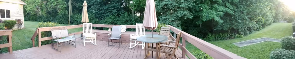 A spacious wooden deck with outdoor furniture including chairs, a bench, and a round table with an umbrella. The deck overlooks a grassy yard bordered by dense green trees and shrubs. A small building is visible on the left side of the image.