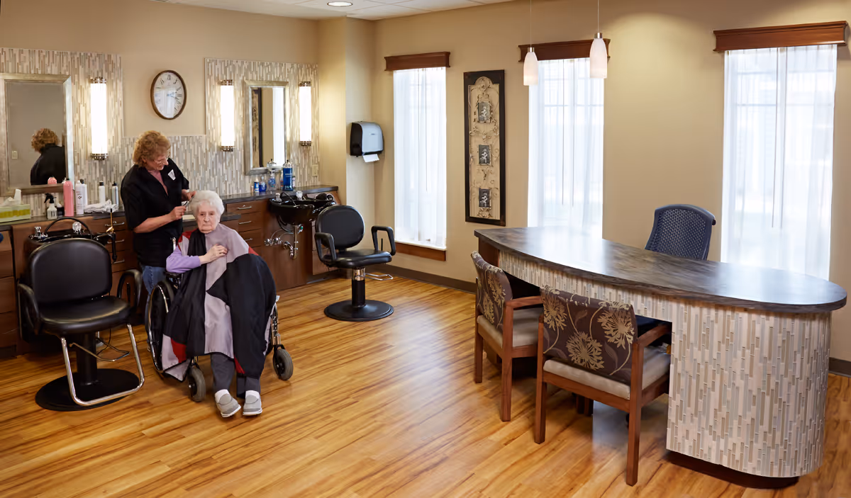 A salon area inside a senior living facility where a stylist tends to an elderly woman in a wheelchair beside salon chairs and a reception desk.
