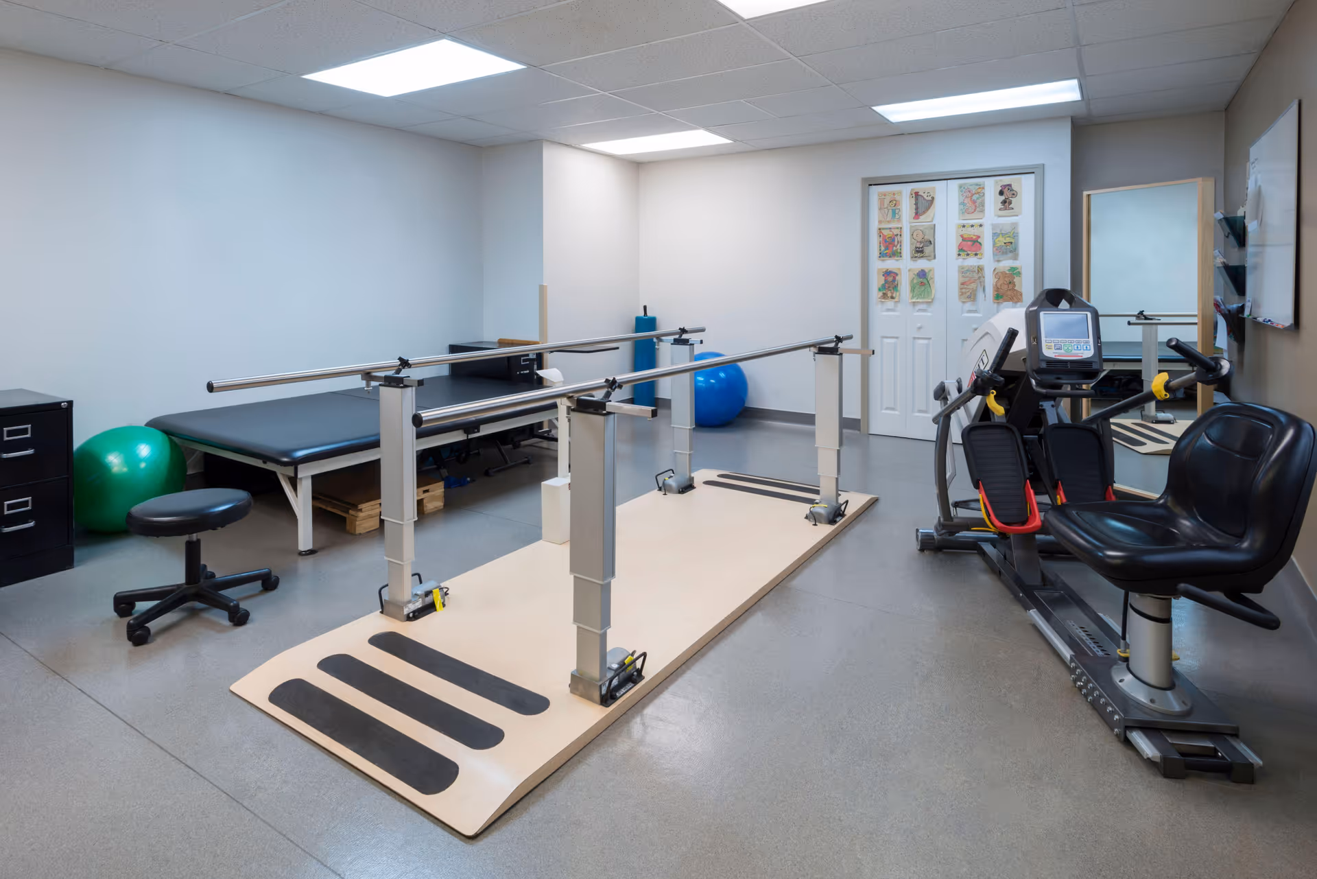A physical therapy or rehabilitation room with parallel bars for walking exercises, an exercise bike, a black padded treatment table, a rolling stool, and exercise balls. The room has a gray floor, white walls, and fluorescent ceiling lights. There are colorful drawings or posters on the door in the background.