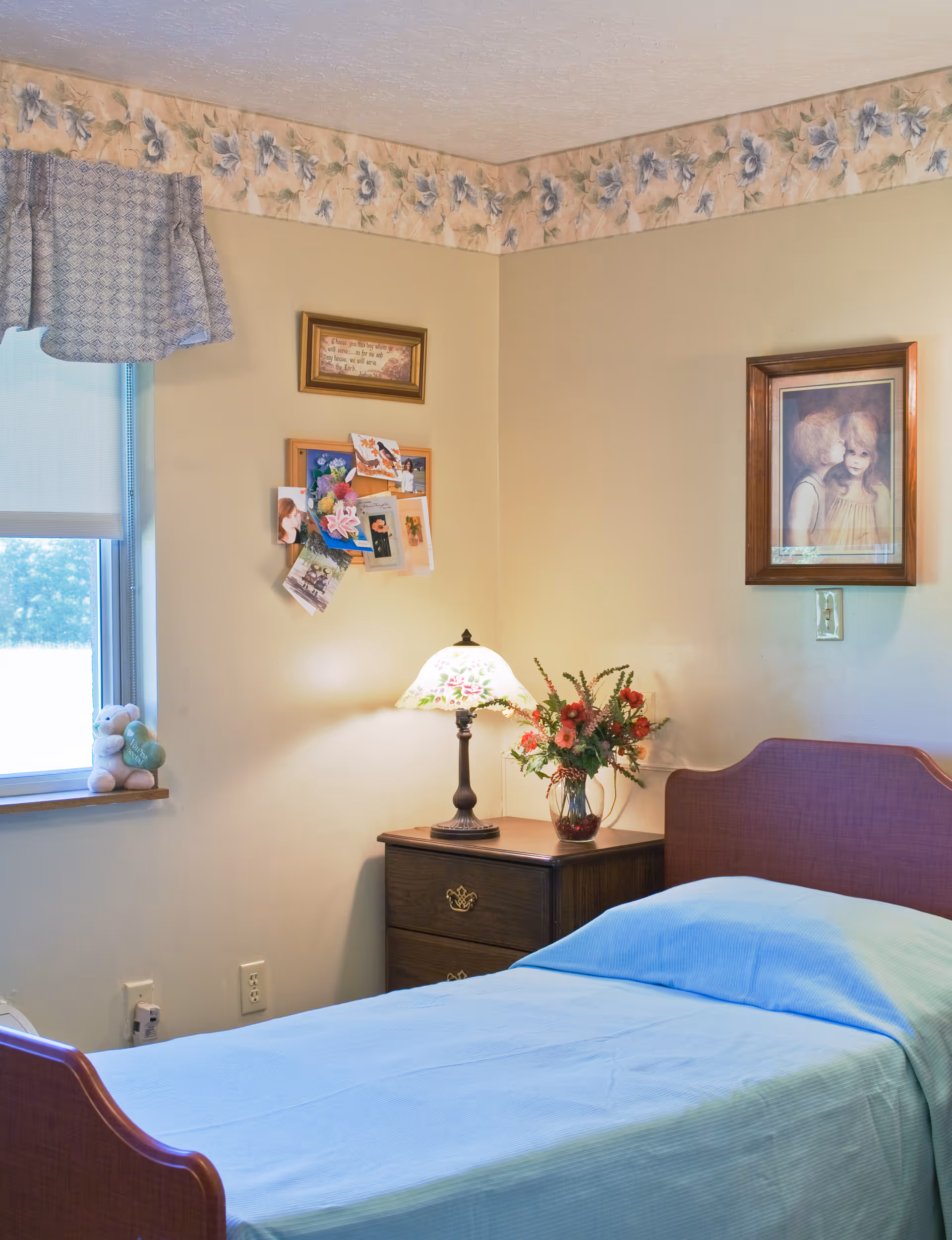 Single-bed bedroom with a nightstand holding a lamp and vase of flowers, wall art and a bulletin board next to a window with a valance.