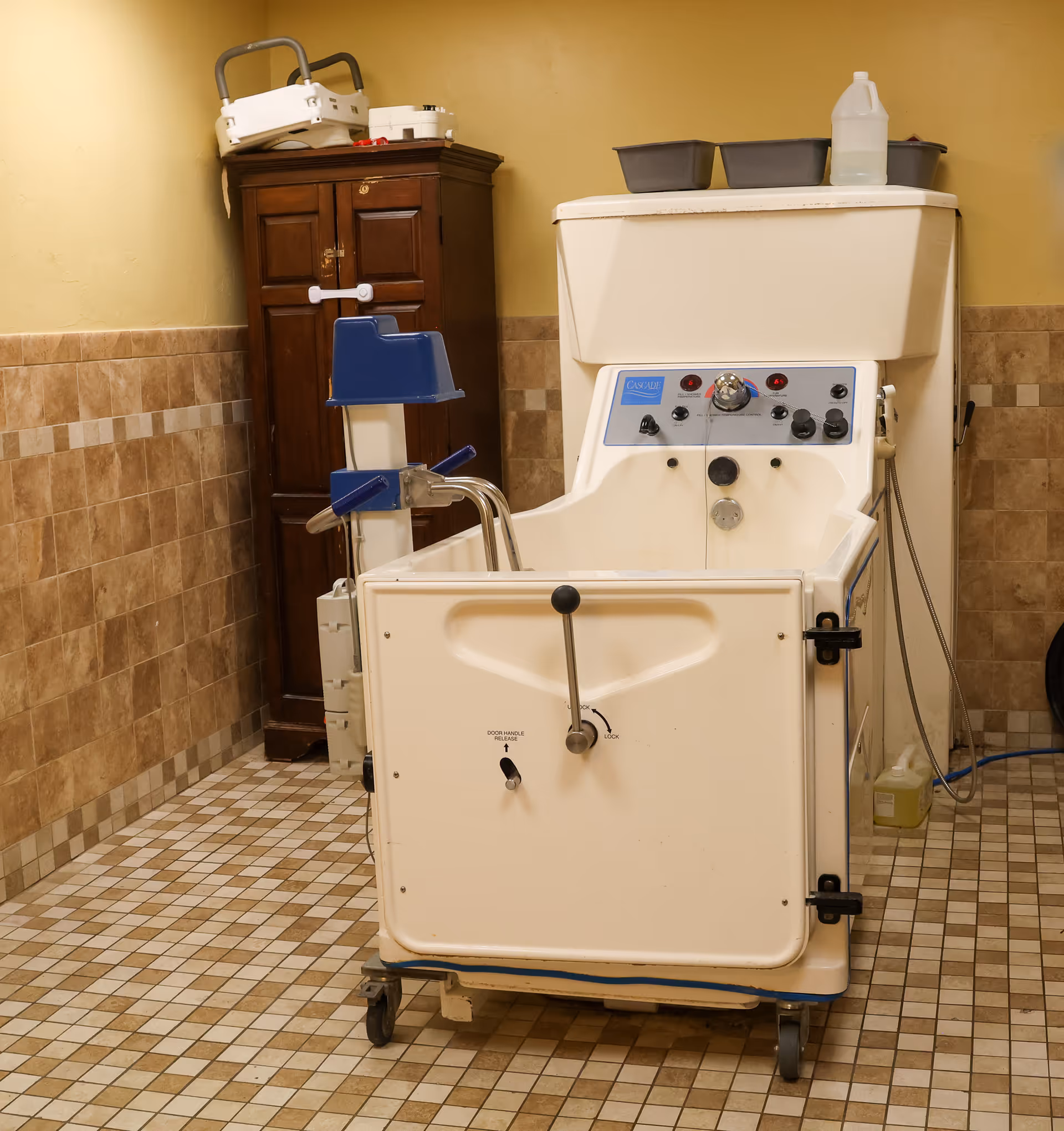 A specialized medical bathtub with control knobs and a door for easy access, situated in a tiled room with beige and brown walls. There is a wooden cabinet in the corner with medical equipment on top and cleaning supplies nearby.