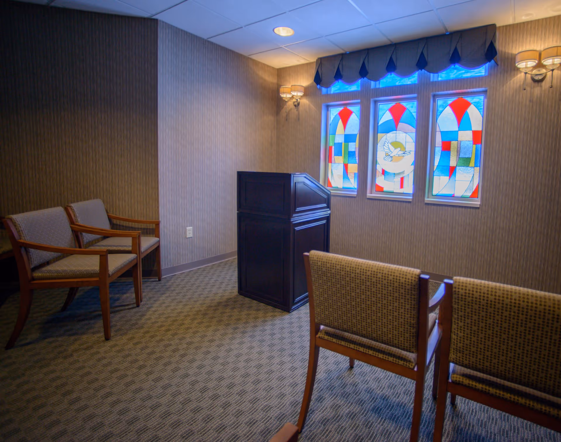 A small room with patterned carpet and wallpaper, featuring a dark wooden podium in the center. There are three stained glass windows with colorful geometric designs and a dove in the middle window. Four wooden chairs with cushioned seats and backs are arranged facing the podium. Two wall-mounted light fixtures with lampshades provide warm lighting.