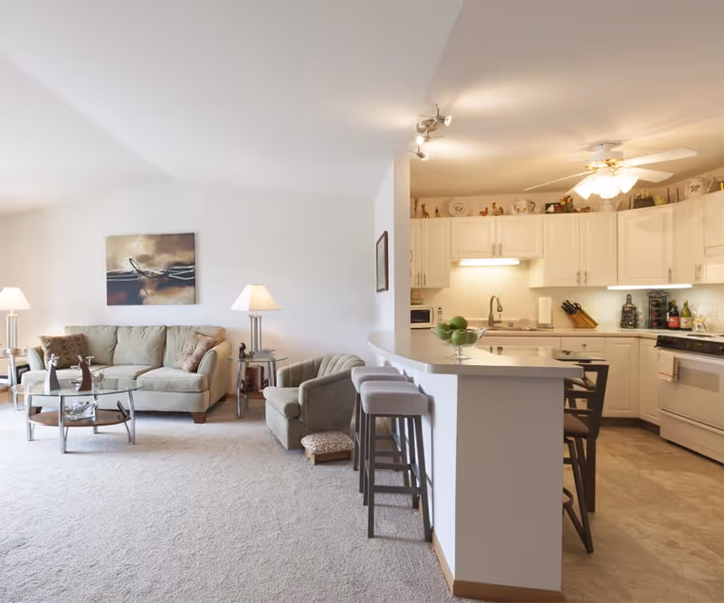 A bright and clean living room and kitchen area in a senior living facility. The living room features a beige sofa, an armchair, two side tables with lamps, a glass coffee table, and a painting on the wall. The kitchen has white cabinets, a ceiling fan with lights, a stove, a microwave, and a counter with three bar stools. The space is carpeted in the living room and has a tiled floor in the kitchen.