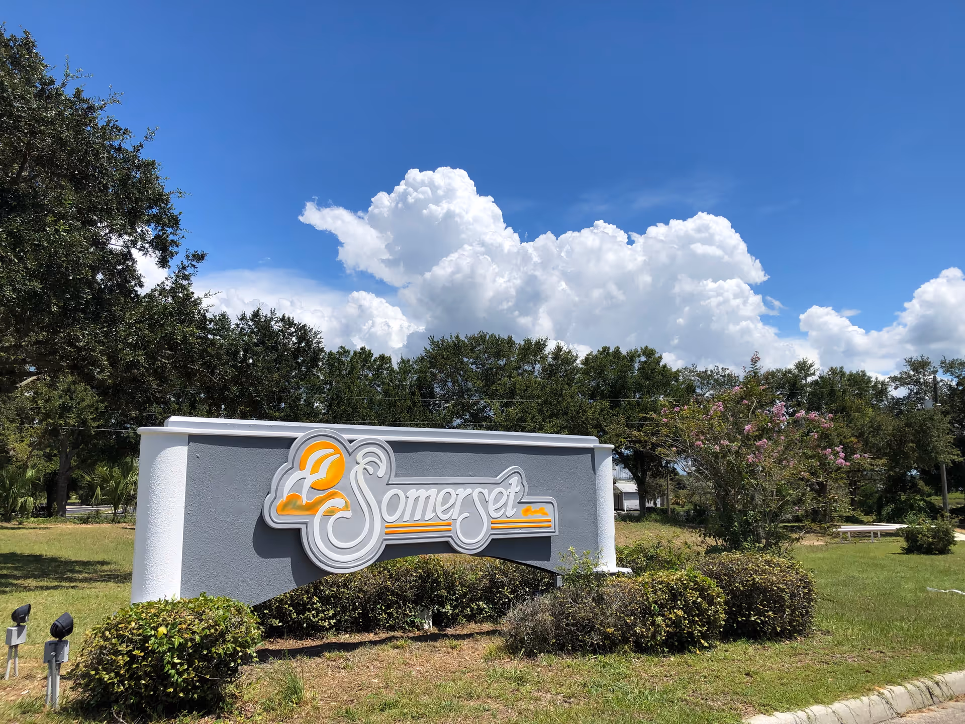 Outdoor view of a Somerset Assisted Living & Memory Care facility sign surrounded by bushes and trees under a blue sky with large white clouds.
