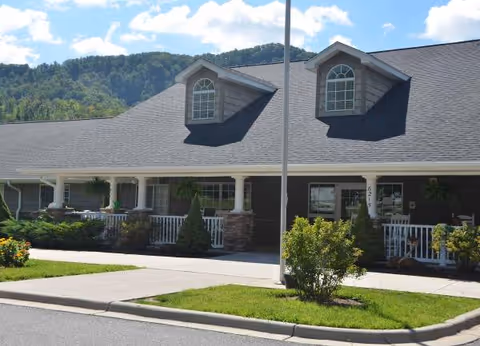 Exterior view of a single-story assisted living facility building with a dark roof, two dormer windows, a covered porch with white railings, and landscaping including bushes and small trees. A flagpole is visible in front of the building, with a backdrop of green wooded hills under a partly cloudy sky.