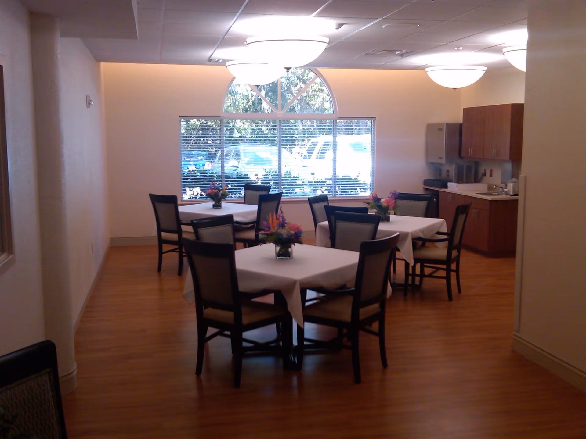 A dining area with four square tables covered with white tablecloths, each surrounded by four chairs. Each table has a small floral centerpiece. The room has wooden flooring, a large window with blinds, and overhead ceiling lights. There is a kitchenette area with cabinets and a countertop on the right side of the room.