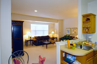 Open-plan living area with a kitchen sink and counter in the foreground and a furnished living room with a sofa, coffee table, and lamps in the background.