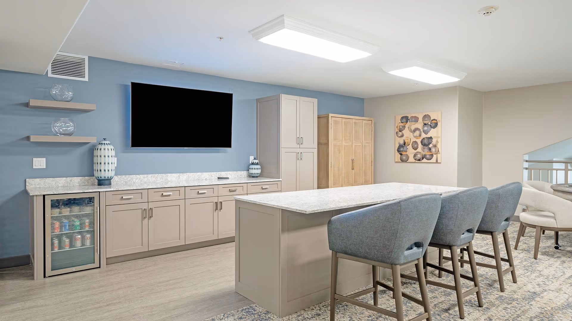 A modern common area with a long kitchen island featuring a marble countertop and three blue upholstered bar stools. Behind the island, there is a wall with beige cabinets, a small refrigerator stocked with drinks, decorative vases, and a large flat-screen TV mounted on a blue accent wall. To the right, there is a wooden cabinet and a piece of abstract wall art. The space has light wood flooring and is well-lit with ceiling lights.