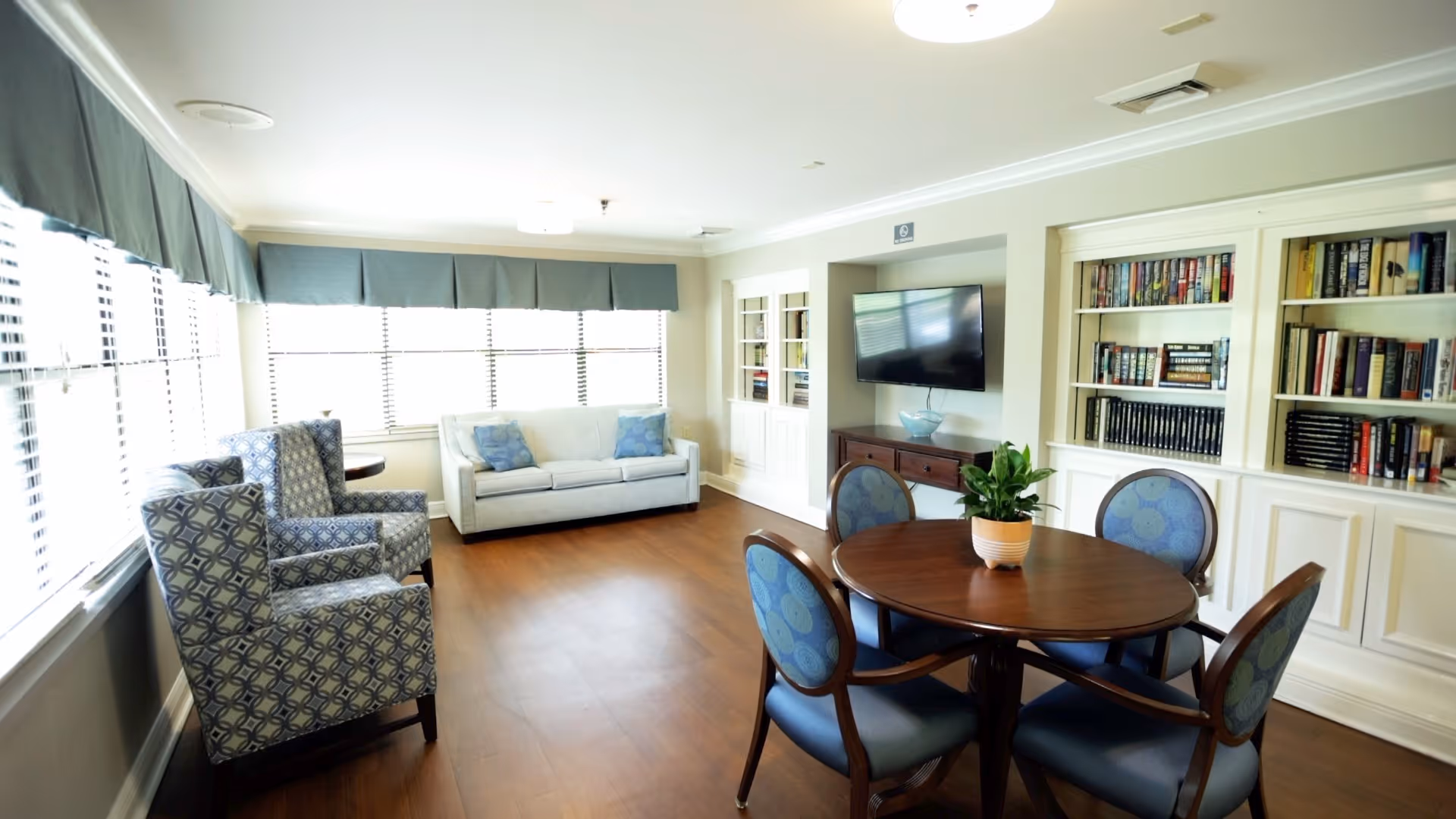Bright communal living room with armchairs, a sofa, round table, bookshelves and a wall-mounted TV.