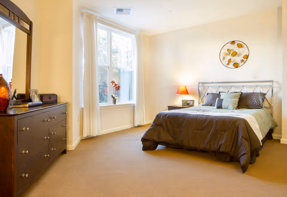 A bright bedroom with a large window covered by white curtains, a bed with a metal headboard and multiple pillows, a nightstand with a red lamp and a framed photo, a wooden dresser with a mirror, decorative items, and a beige carpeted floor.