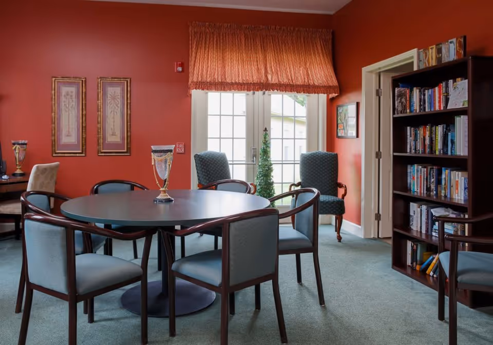 A cozy room with a round table surrounded by six chairs, two armchairs near a window with a red valance, a bookshelf filled with books, and framed artwork on the red walls.