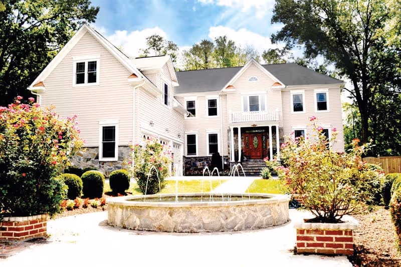 Front exterior view of a large, two-story beige house with multiple windows and a dark roof. The entrance features a red door with glass panels and a small balcony above it. In front of the house is a circular stone fountain with water jets, surrounded by neatly trimmed bushes and flowering plants. Tall trees are visible in the background under a partly cloudy sky.