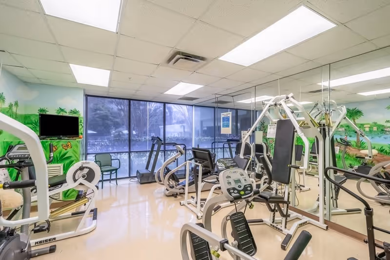 A senior living facility exercise room with various fitness equipment including stationary bikes, treadmills, and weight machines. The room has large windows with a view of greenery outside and walls decorated with a colorful mural of a tropical landscape. There is a TV mounted on the wall and a mirrored wall reflecting the equipment.