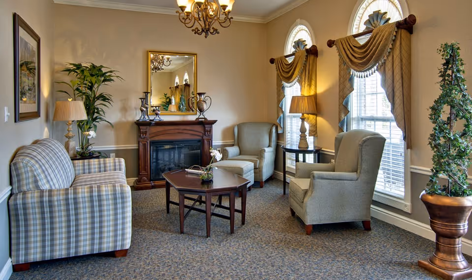 A cozy senior living facility lounge with a plaid sofa, two armchairs, a wooden coffee table, a fireplace with decorative items on the mantel, two windows with draped curtains, table lamps, and potted plants.