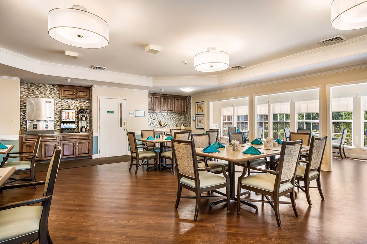 A bright dining room with wooden floors and several tables set with teal napkins. The room features large windows letting in natural light, wooden chairs with cushioned seats, and a beverage station with a coffee maker and ice machine against the wall.