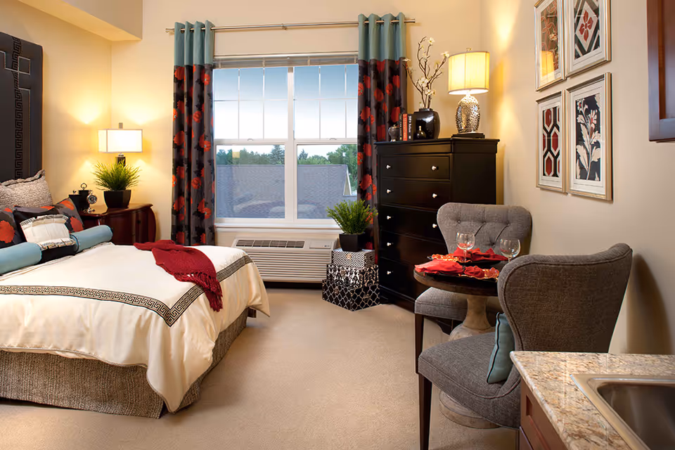A cozy bedroom in an assisted living facility featuring a neatly made bed with decorative pillows and a red throw blanket. The room has a large window with patterned curtains, a black dresser with a lamp and decorative items on top, two upholstered chairs around a small round table set with two wine glasses and red napkins, and framed artwork on the wall. A small kitchenette countertop with a sink is visible in the foreground.