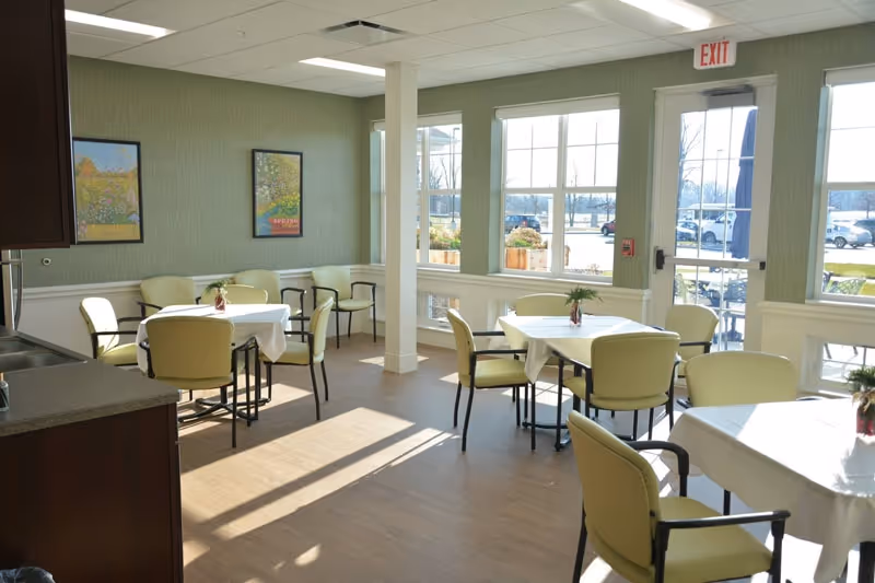 Bright dining room with round tables covered in white cloths, green cushioned chairs, large windows, and framed artwork on the walls.