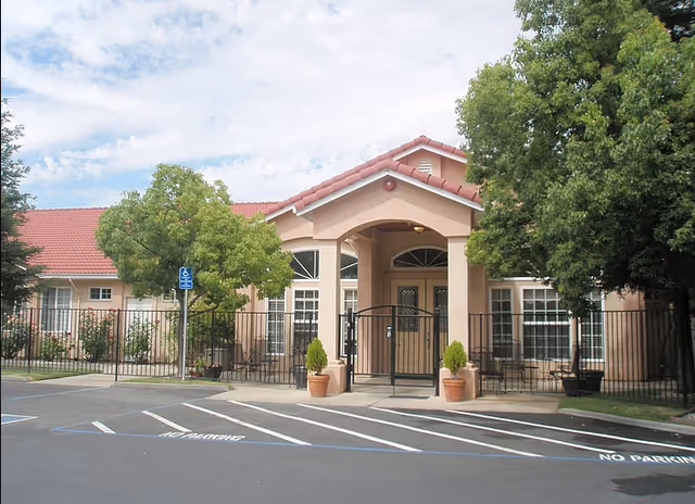 Exterior view of a single-story building with a red-tiled roof and beige walls, featuring a gated entrance with two potted plants on either side. There are trees on both sides of the entrance and a parking lot with marked spaces and a handicapped parking sign in front.