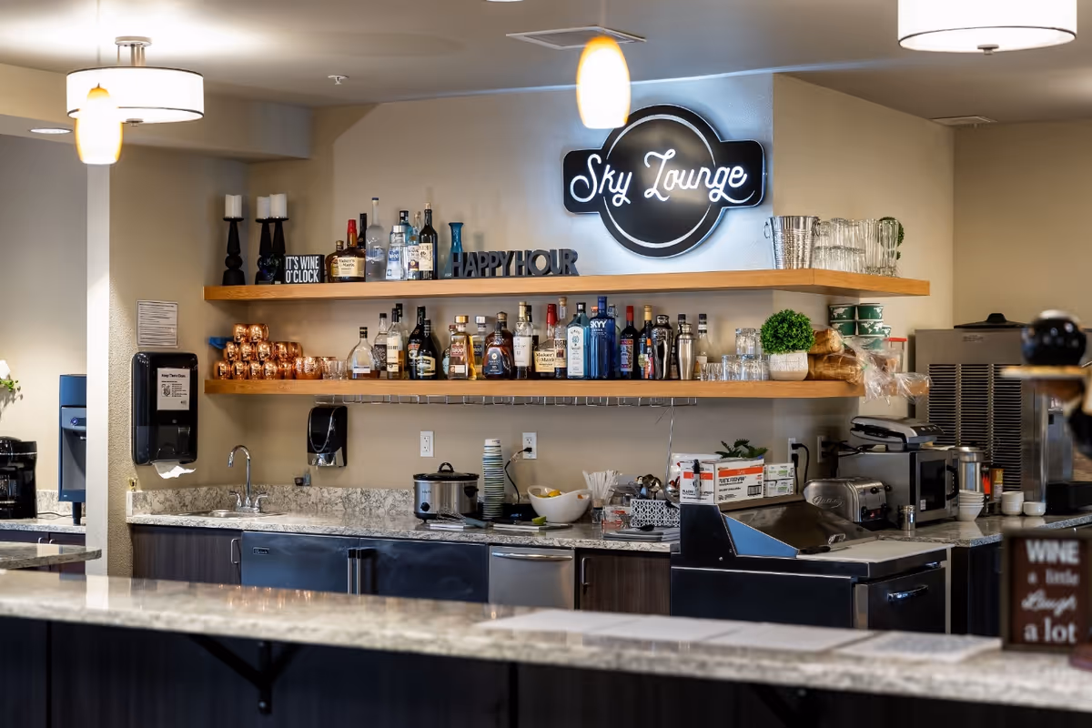 Interior view of a lounge bar area named Sky Lounge with shelves holding various bottles of liquor, glasses, and decorative items. The counter has a granite surface with a sink, coffee maker, and other kitchen appliances visible. The area is well-lit with ceiling lights.