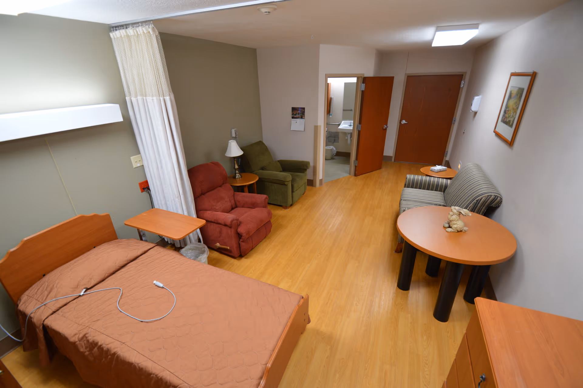 A senior care facility room with a single bed covered in a brown blanket, a red recliner chair, a green armchair, a small round wooden table with a lamp, a striped loveseat, and another round wooden table with a small stuffed animal on it. The room has wooden flooring, light-colored walls, and an open door leading to a bathroom.