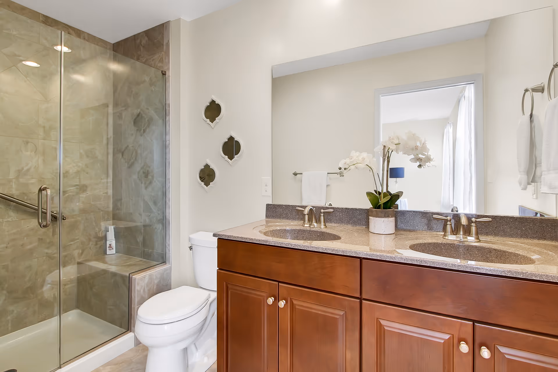 A clean and modern bathroom featuring a glass-enclosed shower with tiled walls, a white toilet, and a double sink vanity with brown wooden cabinets and a granite countertop. A potted orchid is placed between the two sinks, and a large mirror reflects part of an adjacent room. White towels hang on a towel rack and ring.