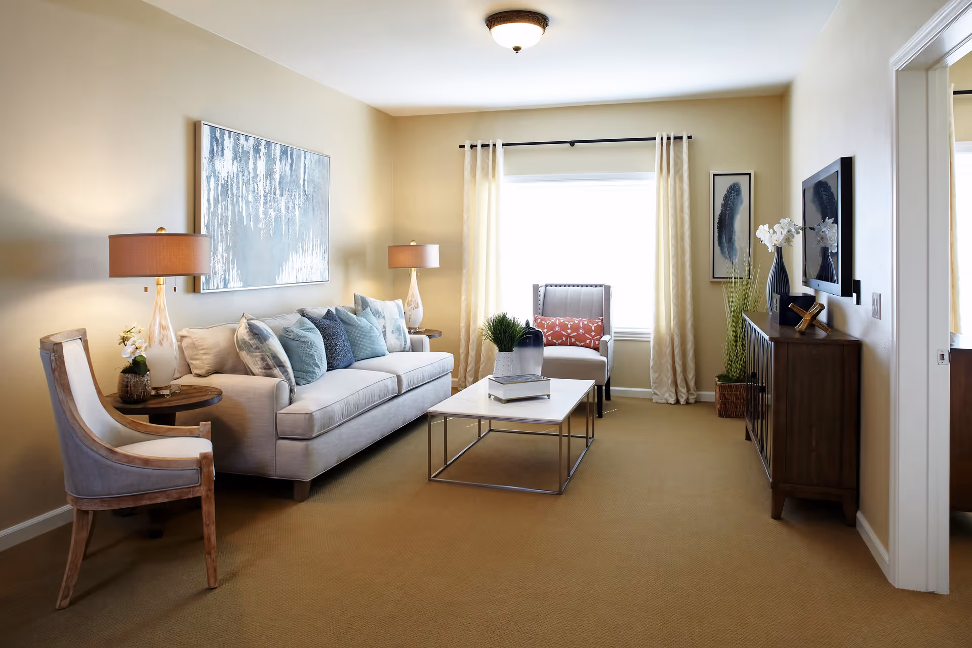 A cozy living room with a beige sofa adorned with multiple blue and gray cushions, a wooden armchair with light upholstery, two side tables each with a lamp, a rectangular coffee table with books and a plant, a wooden cabinet with decorative items, and a window with light curtains letting in natural light.