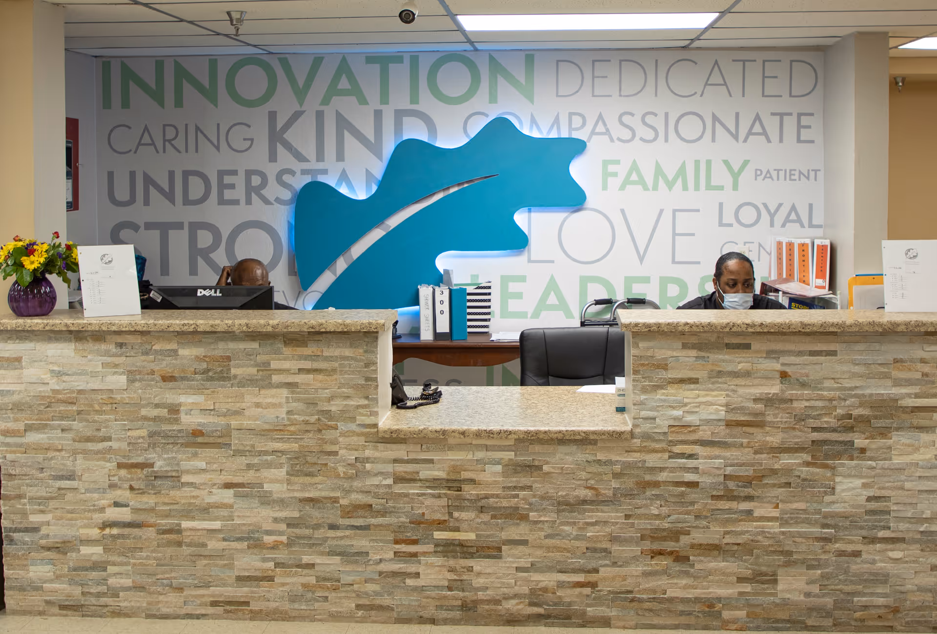Reception desk inside a care facility with a stone-front counter, large decorative wall art and a masked staff member working behind the desk.
