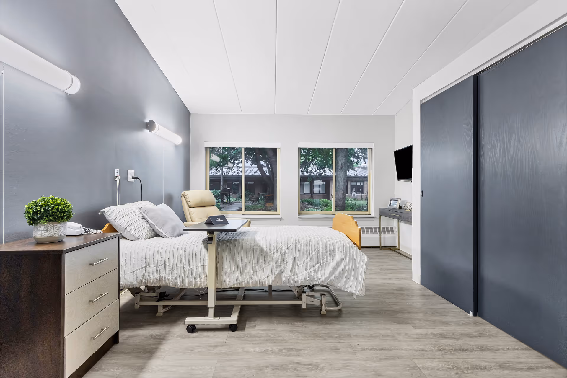 A bright and clean bedroom in a senior living facility featuring a hospital-style bed with white bedding, a beige recliner chair, a small overbed table, a dark wood nightstand with a potted plant and telephone, two large windows showing an outdoor garden view, a wall-mounted TV, and a desk with a chair. The room has light wood flooring, a gray accent wall, and sliding closet doors.