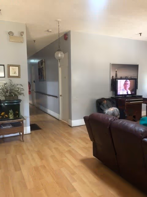 Interior view of a senior living facility showing a hallway with handrails, a small table with a fish tank and plant, a brown leather couch, and a television displaying a man in a suit. The walls are light gray and the floor is wooden.