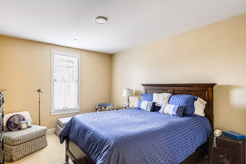Sunlit bedroom with a large bed dressed in blue bedding, a wooden headboard, nightstands, and a window with blinds.