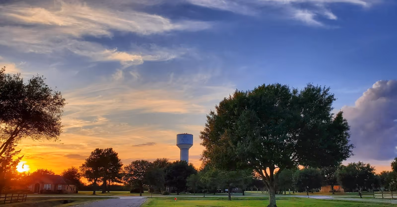A scenic outdoor view at sunset with a partly cloudy sky, a water tower labeled 'Biscoe Falls', trees, a road, and houses in the background.