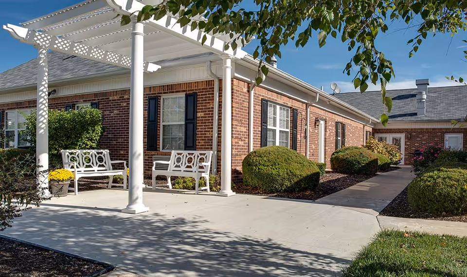 Outdoor seating area at Chestnut Glen Senior Living featuring white benches under a white pergola, surrounded by neatly trimmed bushes and a brick building with black shutters in the background under a clear blue sky.
