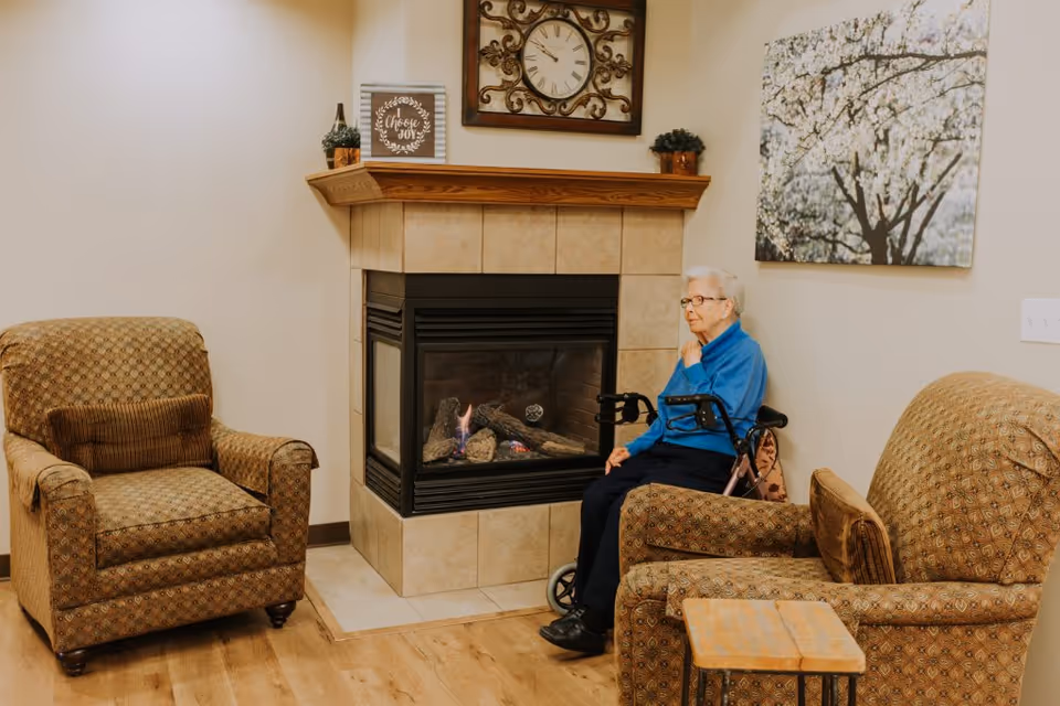 An elderly woman sitting in a wheelchair near a corner fireplace in a cozy living room area with two patterned armchairs, a small wooden side table, a wall clock above the fireplace, and a large floral painting on the wall.