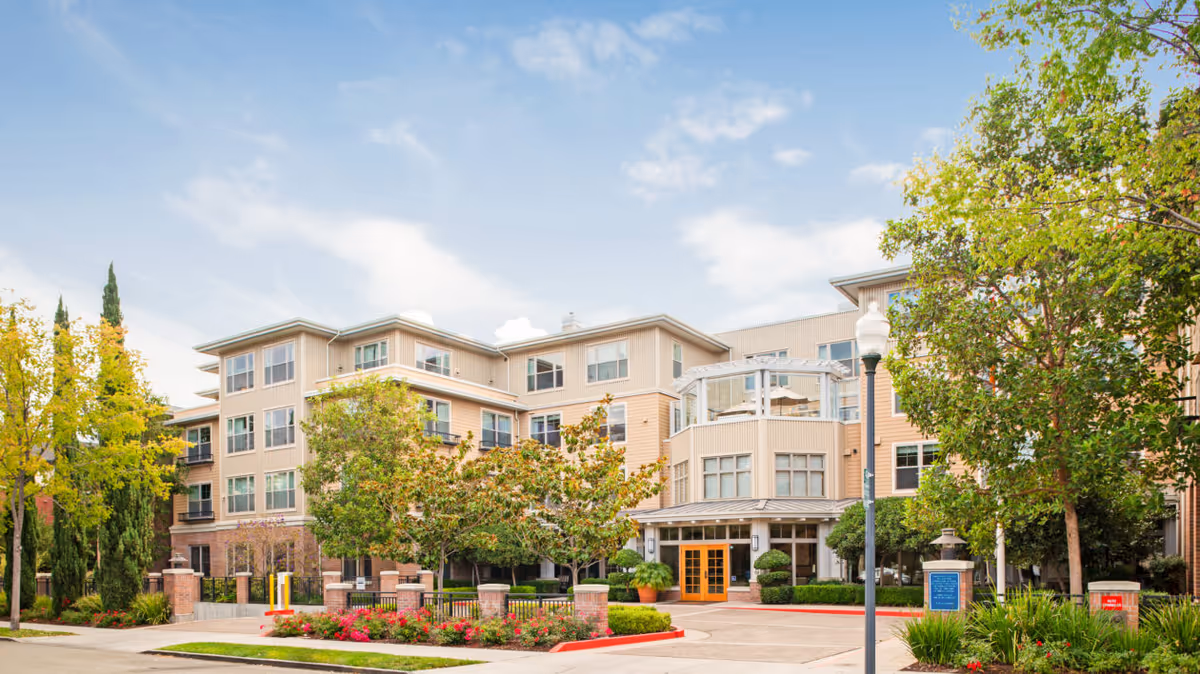 Four-story beige senior living building with a landscaped entrance, trees, and clear blue sky.