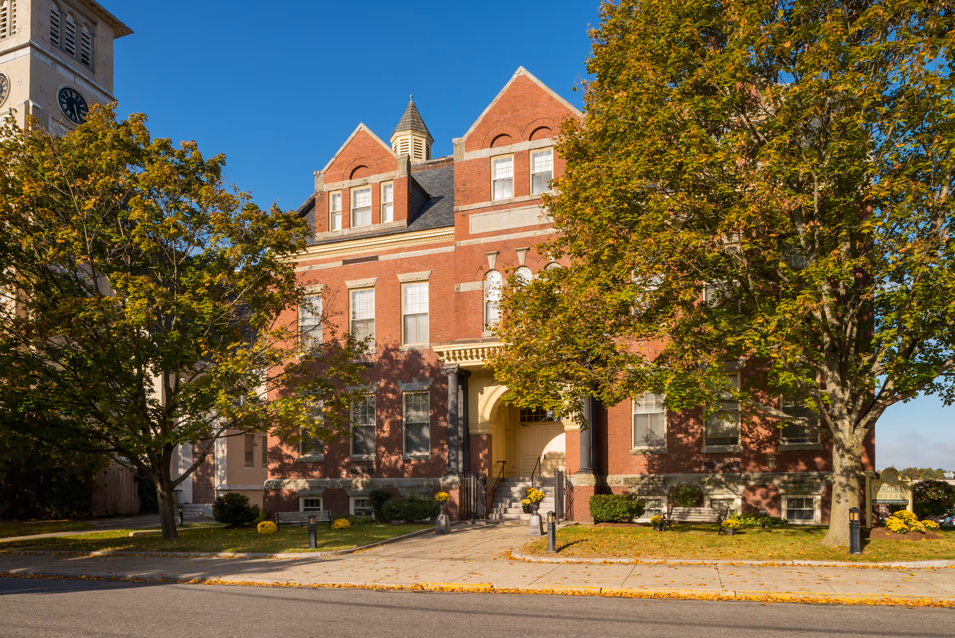 A large, historic red brick building with multiple windows and a central entrance with steps leading up to it. The building is surrounded by trees with green and yellow leaves, indicating early autumn. The sky is clear and blue. There is a sign partially visible on the right side of the image that reads 'Corcoran House'.