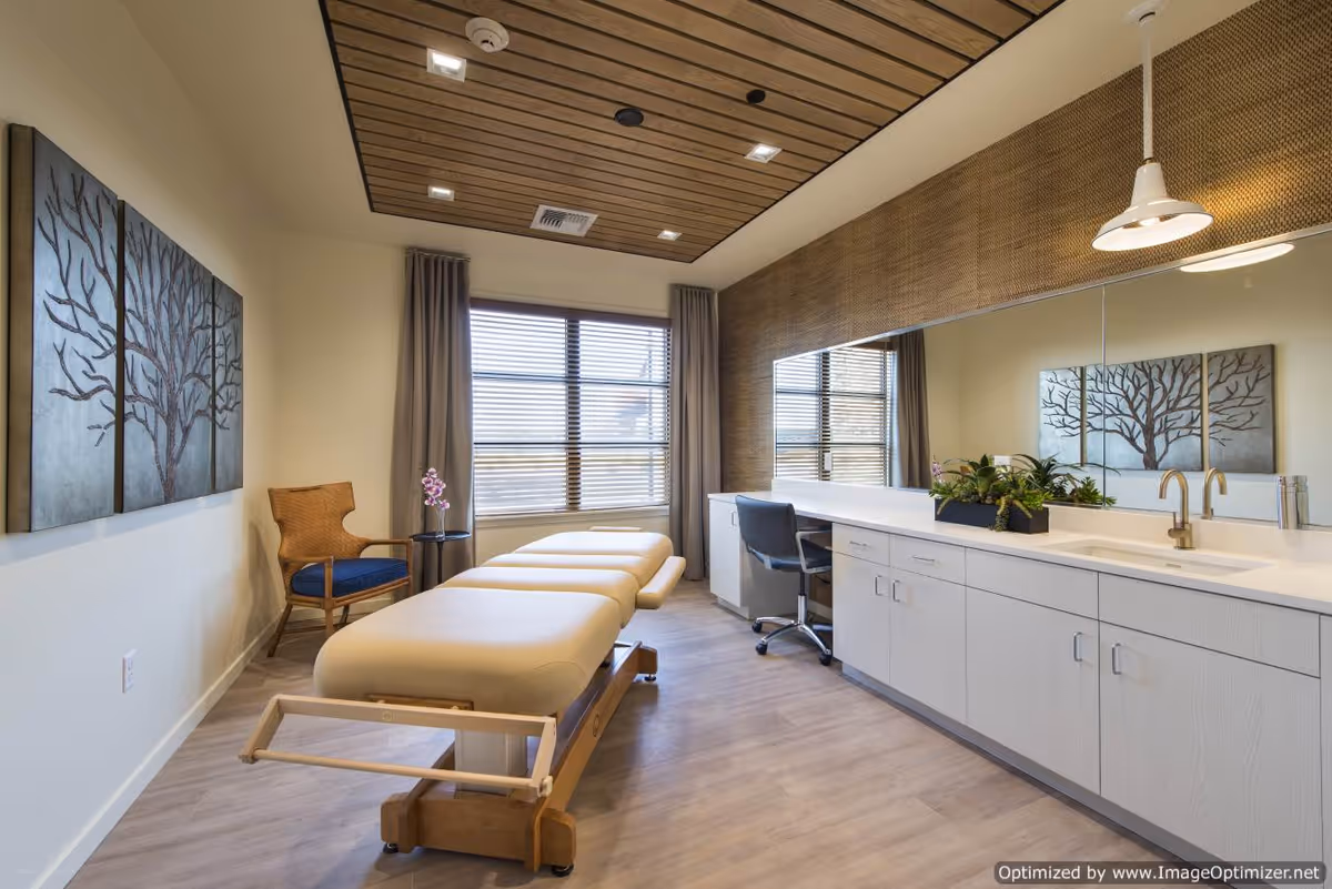 A bright, modern therapy or treatment room with a padded treatment table in the center, a wooden chair with a blue cushion and a small side table with a pink orchid near the window. The room features a large mirror above a long white cabinet with a sink and faucet, a wooden slatted ceiling panel with recessed lighting, and a three-panel artwork of a tree on the wall.