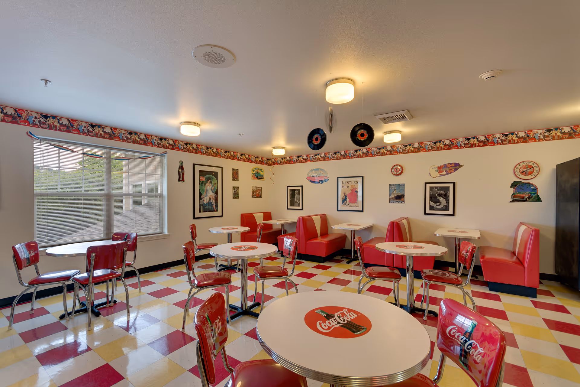 A retro-themed dining room with red and white Coca-Cola branded tables and chairs. The floor has a checkered pattern in red, yellow, and white. Red cushioned booths line the walls, which are decorated with vintage Coca-Cola and other nostalgic posters and signs. Two vinyl records hang from the ceiling. A large window lets in natural light.