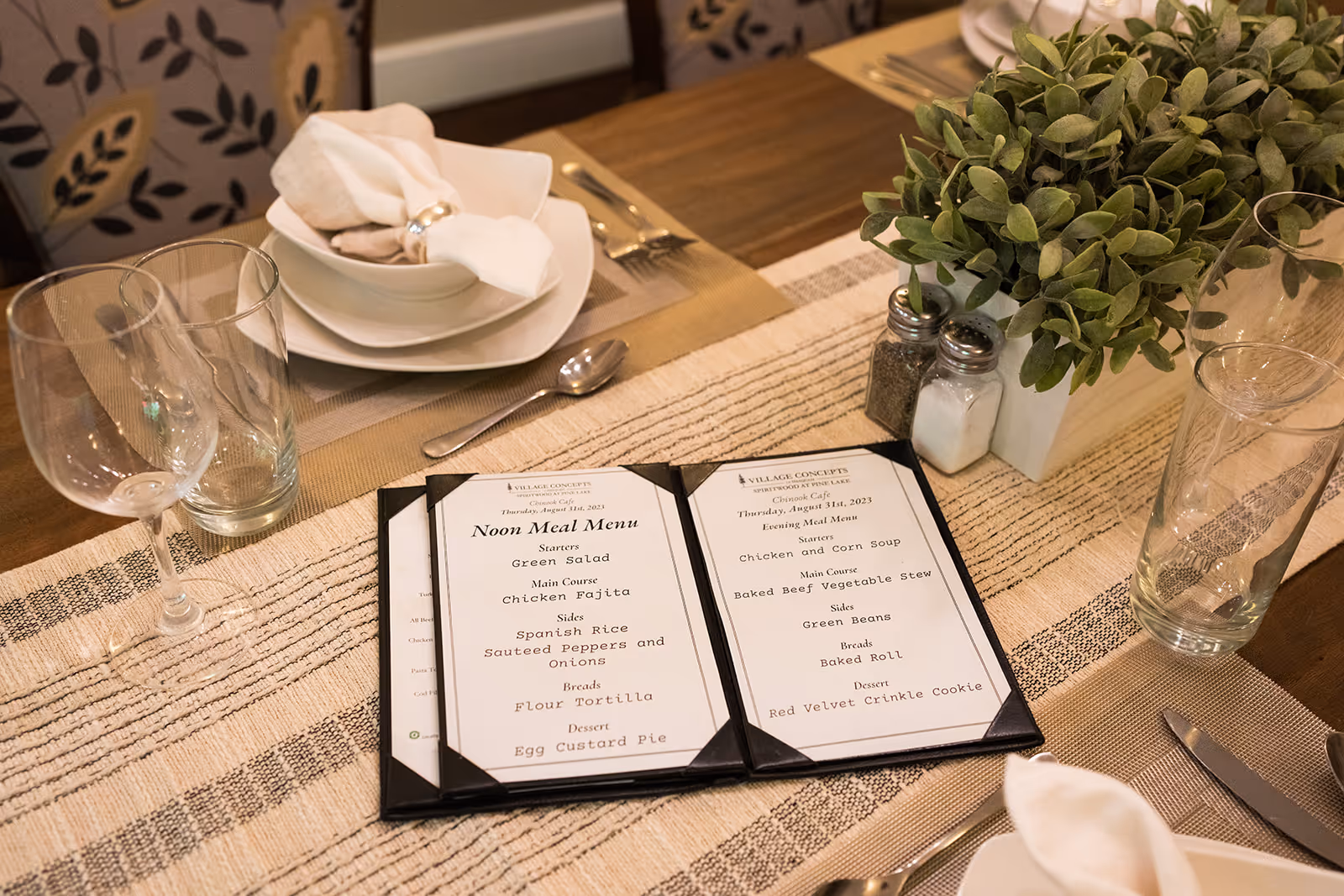 A dining table set with plates, bowls, napkins, silverware, wine glasses, and water glasses. In the center of the table is a small green potted plant and salt and pepper shakers. Two menus from Village Concepts of Issaquah-Spiritwood at Pine Lake are placed on the table, showing the Noon Meal Menu and Evening Meal Menu.