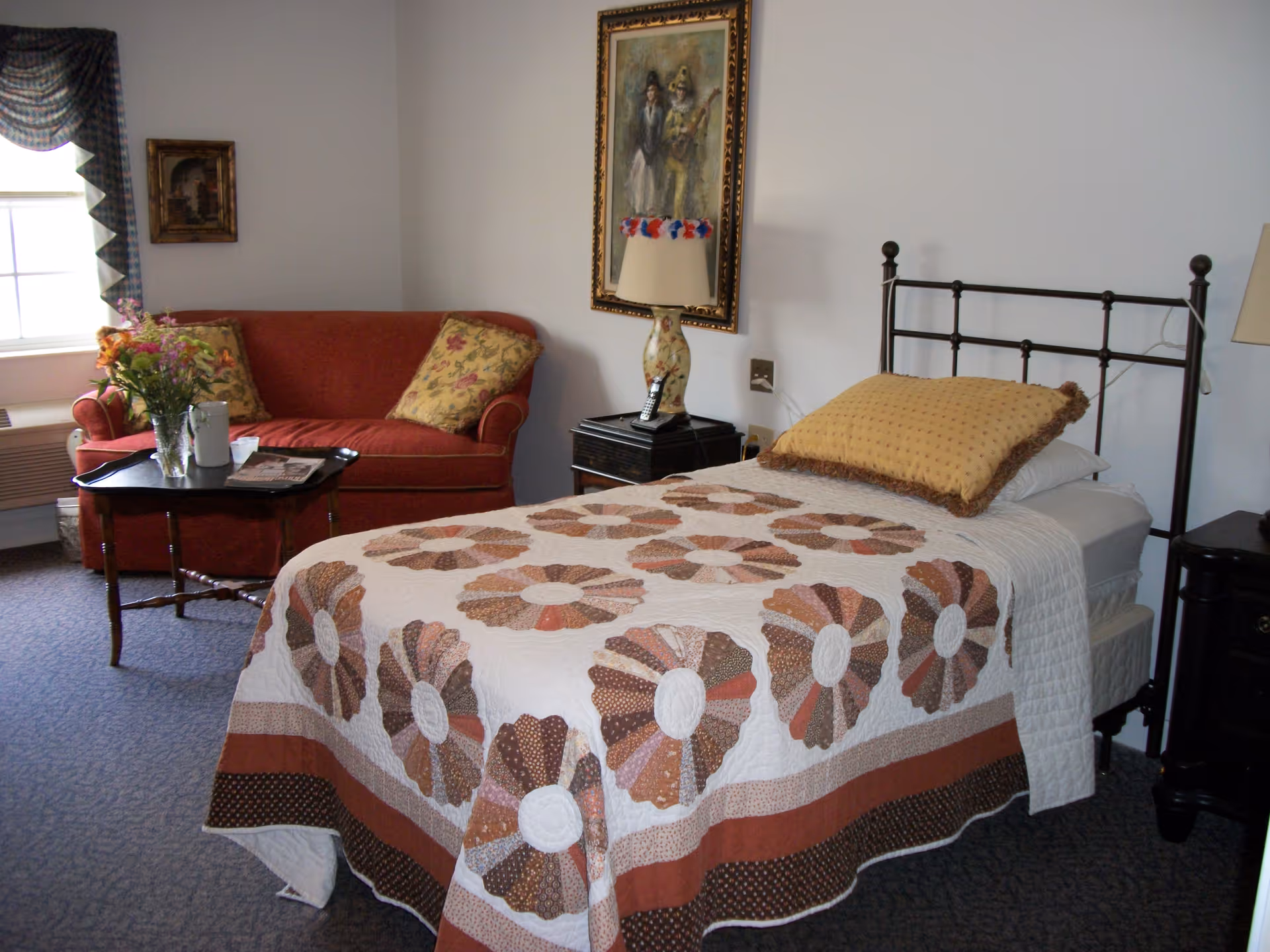 A cozy bedroom with a single metal bed dressed in a patterned quilt, a red loveseat and coffee table, and bedside tables with lamps.
