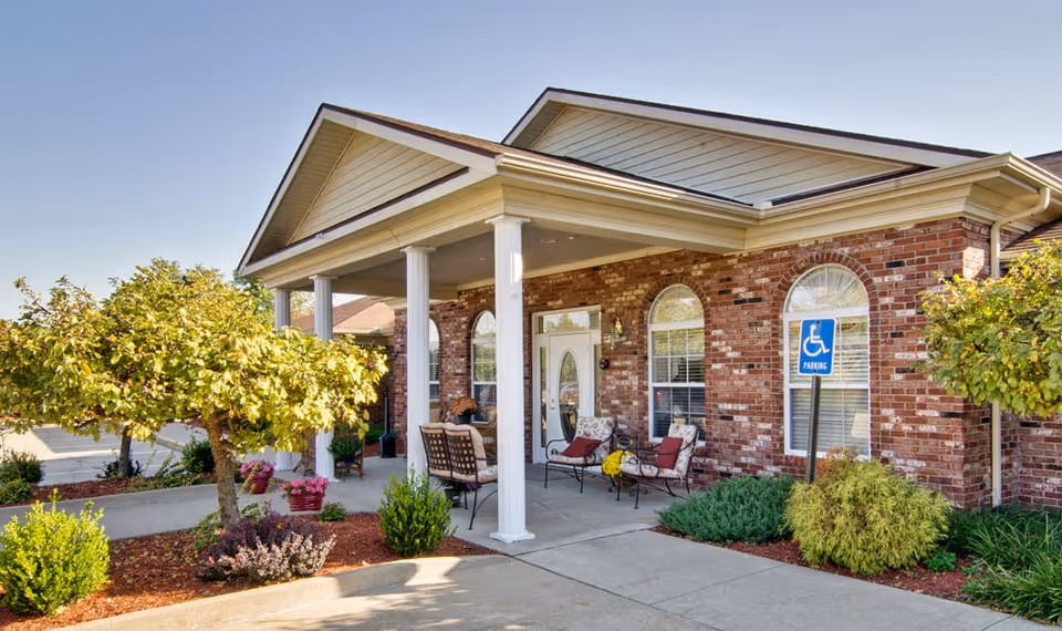 Exterior view of a senior living facility with a brick facade, white columns supporting a covered entrance, outdoor seating with cushioned chairs, landscaped bushes and small trees, and a handicapped parking sign near the entrance.