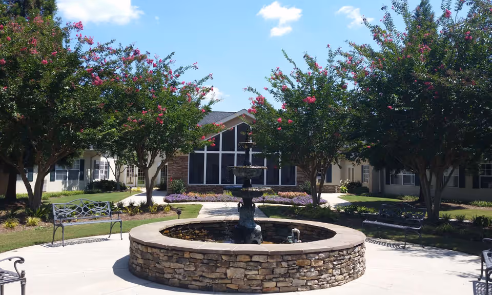 Outdoor courtyard area at Azalea Estates of Fayetteville featuring a circular stone fountain in the center, surrounded by benches and flowering trees under a clear blue sky.