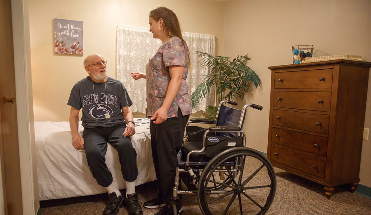 A caregiver speaks with an elderly man seated on a bed in a furnished senior living bedroom with a wheelchair and dresser.