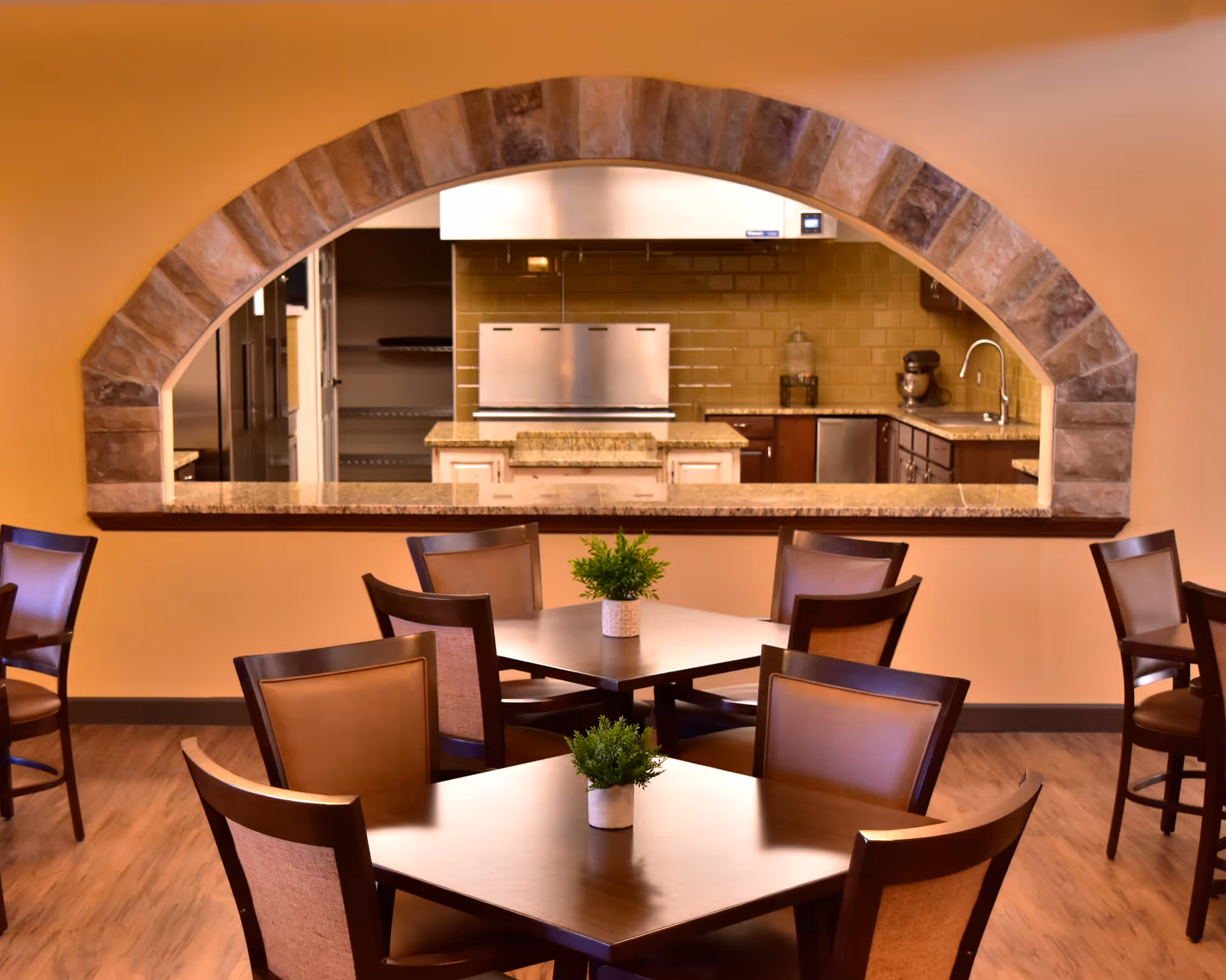 Dining area with wooden tables and chairs, each table decorated with a small green plant. In the background, a kitchen is visible through a stone archway, featuring countertops, a sink, and kitchen appliances.