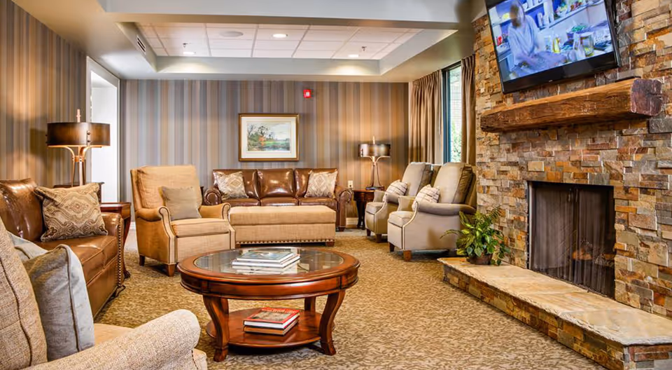 A cozy living room with a stone fireplace on the right side, a flat-screen TV mounted above it, and a variety of seating including leather and fabric armchairs, a leather sofa, and an upholstered ottoman. The room has patterned wallpaper, carpeted floor, two standing lamps, a round wooden coffee table with books, and a framed painting on the back wall.