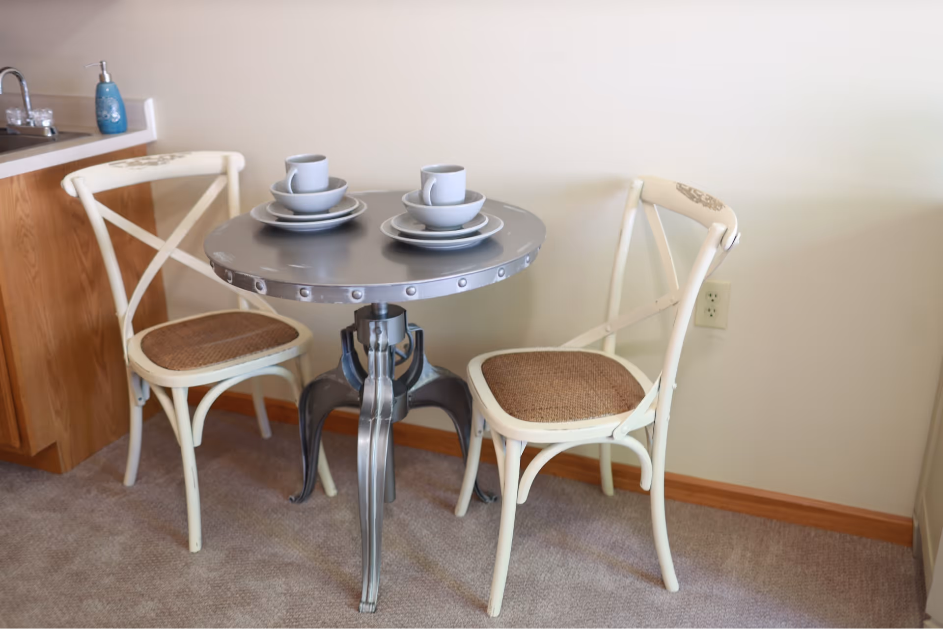 A small round metal table set with two place settings, each including a cup, saucer, and plate, accompanied by two white wooden chairs with woven seats, situated next to a kitchen counter with a sink and soap dispenser.