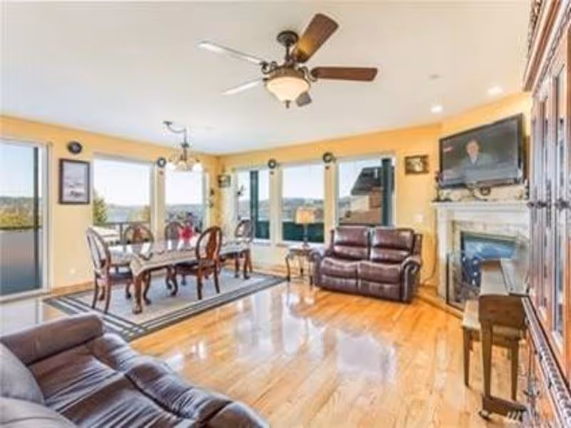 A bright and spacious living room and dining area with wooden floors, yellow walls, and large windows letting in natural light. The dining area features a wooden table with six chairs on a rug, and the living area has two brown leather sofas, a fireplace with a mounted TV above it, and a ceiling fan with lights.