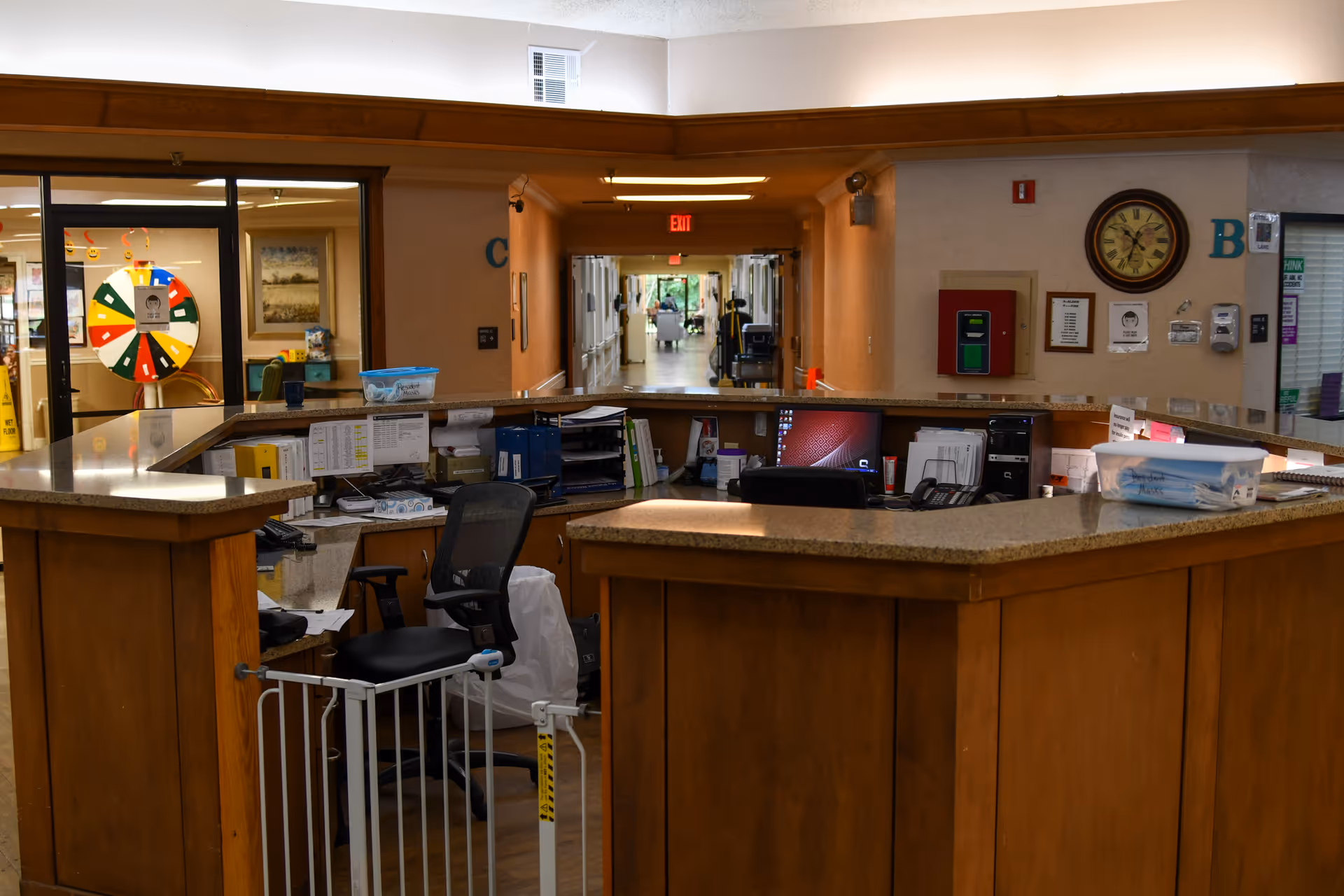 Reception desk area inside Oaks Healthcare Center with a wooden counter, office chair, computer monitors, and various office supplies. In the background, a hallway extends with a person visible at the far end, and a colorful wheel is seen through a glass door on the left.