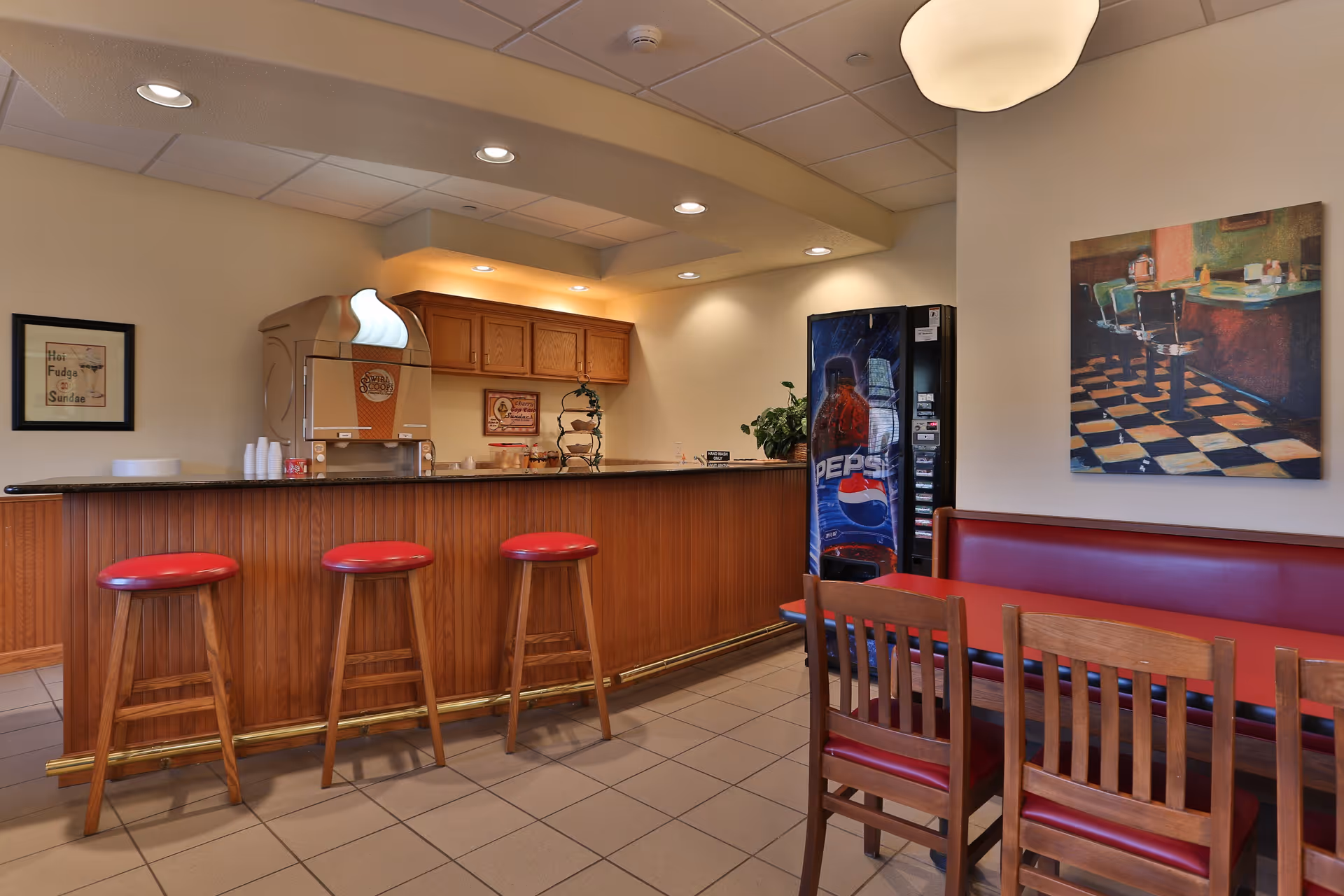 Interior view of a dining area with a wooden counter featuring three red cushioned bar stools, a soda fountain machine, and a Pepsi vending machine. There is a red cushioned bench along the wall with wooden chairs and a table. The walls are decorated with framed artwork and a painting of a diner scene.